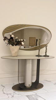 An oval console table with a light marble-look top and pedestal base, paired with a large matching oval wall mirror featuring a wavy gold-toned border. The table is styled with a vase of dried flowers and two decorative candlesticks against a cream wall and marble floor.