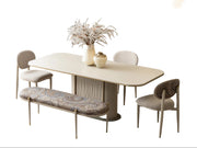 A modern dining room set featuring a light beige oval table with a textured pedestal base, four upholstered dining chairs in neutral tones, a matching patterned upholstered bench, and decorative ceramic vases on the table.