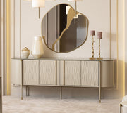 A luxury modern dining room featuring a light wood dining table with a matching upholstered bench and chair, a round backless sideboard with a large oval mirror, a gray armchair, and a small round side table. The room has elegant beige walls with gold paneling, a large window on the left, and three white drum pendant lights hanging above the console.