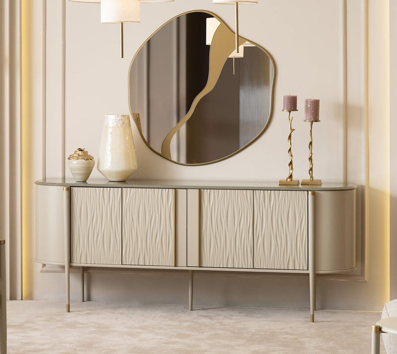 A luxury modern dining room featuring a light wood dining table with a matching upholstered bench and chair, a round backless sideboard with a large oval mirror, a gray armchair, and a small round side table. The room has elegant beige walls with gold paneling, a large window on the left, and three white drum pendant lights hanging above the console.
