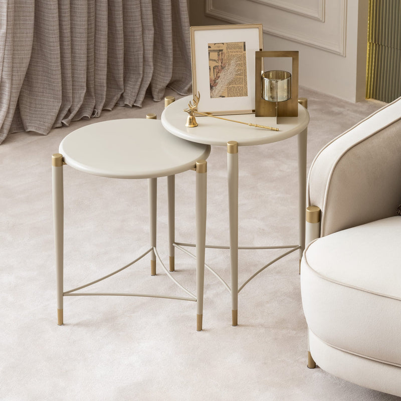 A styled interior scene showing a cream upholstered sofa with two decorative pillows, two small round nesting side tables with gold metal frames, curtains, and floor lamps in a softly lit room.